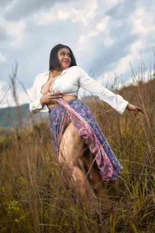 Smiling woman in a white blouse and patterned maxi skirt posing in a dreamy grassy field under a cloudy sky.
