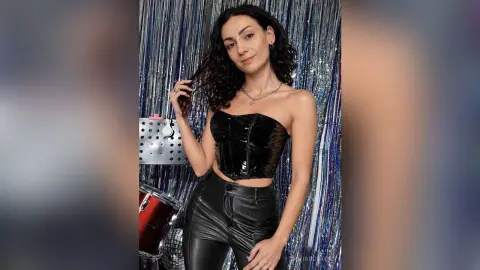 Smiling woman in black leather corset and pants posing against a sparkly silver fringe backdrop.