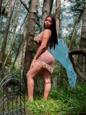 Young woman in a tan fringe mini dress posing in a lush green forest setting with a dreamy mood.