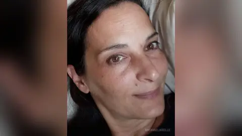 MichaelaBelle smiles closely at the camera with her dark hair framing her face while lying against a white pillow.