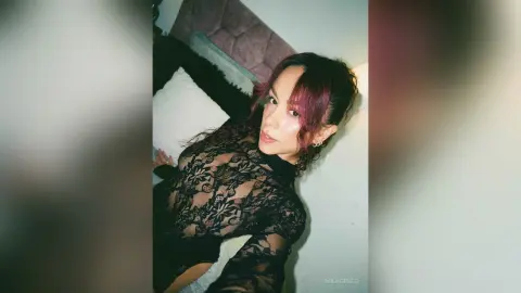 MilaGreco poses in a black lace long-sleeve bodysuit with her dark purple-tinted hair, lying back against a soft headboard.