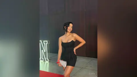 Alison poses in a black strapless mini dress with her hand on her hip against a dark wall.