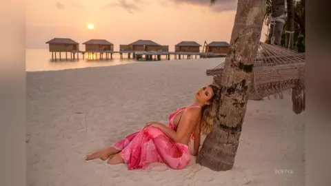 Woman in a pink summer dress relaxing on a tropical beach by a palm tree at sunset.