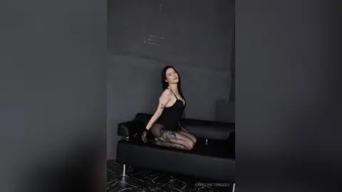 Elegant woman in a black bodysuit sitting on a modern sofa in a moody, dark studio setting.