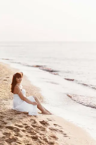 Woman in a white summer dress sitting on a sandy beach by the ocean during a peaceful sunset.