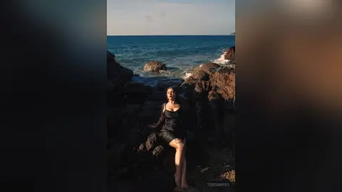 Elegant woman in a black dress standing on dark coastal rocks by the ocean at sunset.