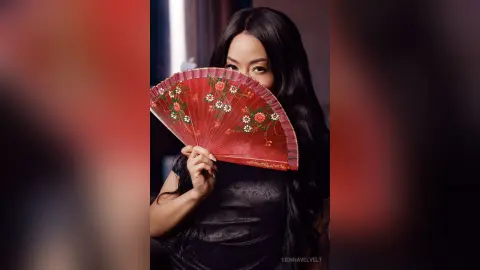 Elegant woman in a black dress holding a red floral folding fan in a moody, cinematic indoor setting.