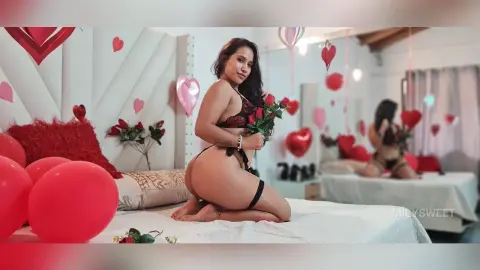 Curvy woman in black lace lingerie kneeling on a bed surrounded by red heart balloons in a romantic bedroom.