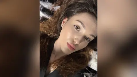 Young woman with wavy brown hair wearing a black top looking thoughtfully at the camera in soft indoor lighting.