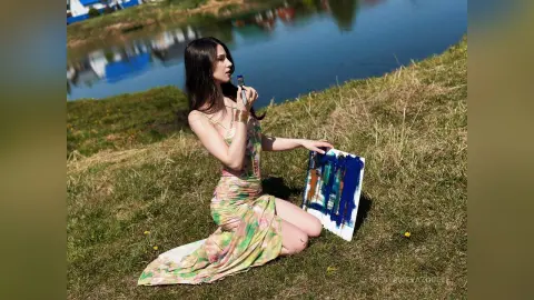 Young woman in a floral sundress painting on canvas by a sunny riverbank.