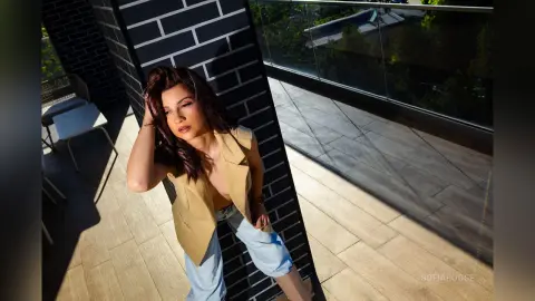 SofiaRoose poses on a sunny balcony wearing an open tan vest and light blue pants, running her hand through her long dark hair.