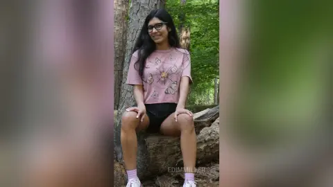 Smiling young woman in glasses and pink graphic t-shirt sitting on a log in a lush green forest.