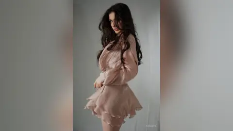 Elegant woman in a flowing blush pink satin dress posing against a minimalist grey background with a dreamy mood.