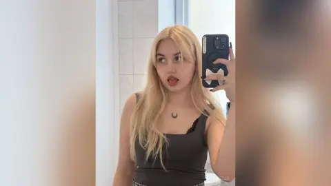 LillyEyes poses for a mirror selfie wearing a black tank top, showing off her long blonde hair and a crescent moon necklace.