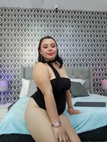 Curvy woman in a black bodysuit sitting on a bed with patterned wallpaper in a cozy bedroom setting.