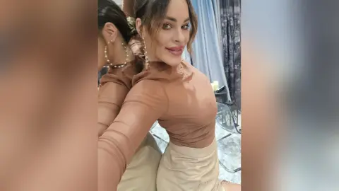 Smiling woman in a brown long-sleeve top and beige trousers posing for a mirror selfie in a bright room.