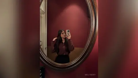 Young woman in a brown sweater taking a moody mirror selfie in a red room.