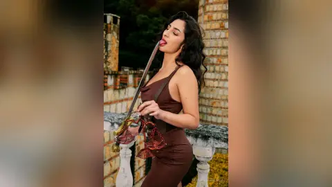 VeronicaValdez licks a long metal rod while wearing a brown sleeveless dress against a stone balcony background.