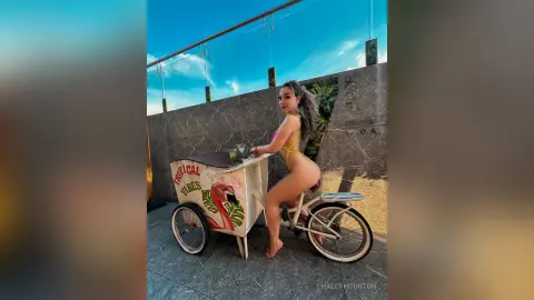 Young woman in a yellow bikini posing with a vintage tropical drink cart on a sunny rooftop terrace.