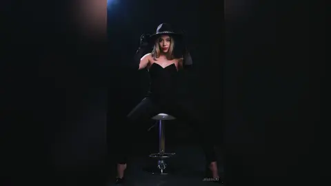 JessieLiu wears a black strapless bodysuit and a black hat while sitting on a white stool against a dark background.