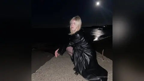 Blonde woman in a black leather trench coat sitting by the ocean under a full moon at night.