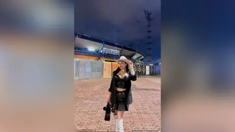Stylish woman in a black crop top, mini skirt, and white boots posing at a stadium under night lights.
