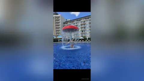 Young woman in a bikini enjoying a refreshing water feature at a luxury hotel pool on a sunny day.
