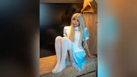 Blonde cosplayer in a blue and white maid outfit sitting on a wooden surface in a moody, dark room.