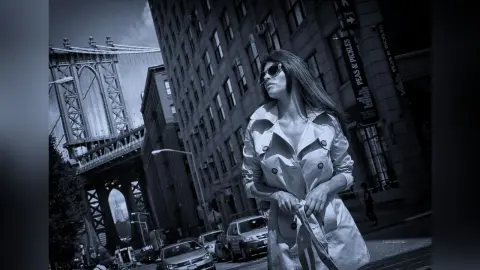 Stylish woman in a trench coat and sunglasses walking on a city street near the Manhattan Bridge.