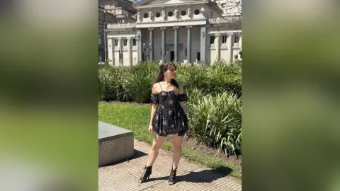 Young woman in a black floral mini dress posing in a sunny urban park near classical architecture.