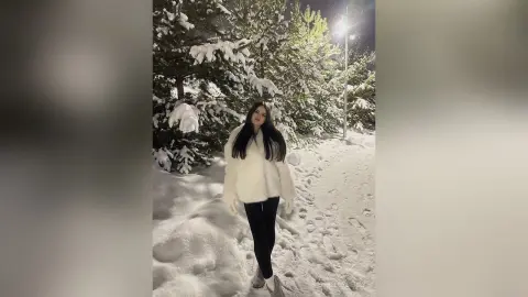 Woman in a white faux fur coat standing in a snowy pine forest at night under soft moonlight.