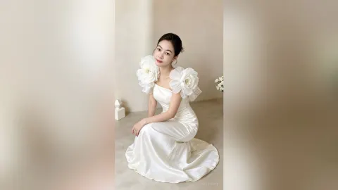CelesteQi — CelesteQi poses in a white satin gown with large floral shoulder accents while kneeling on the floor.