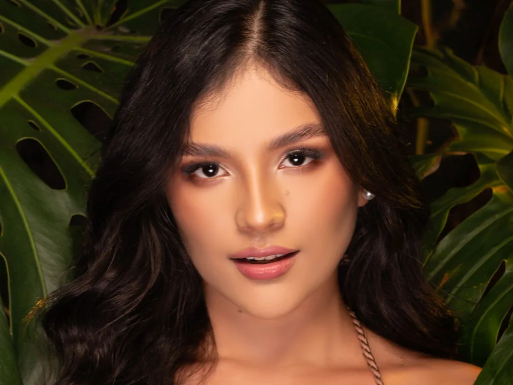 MiahRivers poses with long wavy dark hair against a background of large green tropical leaves.