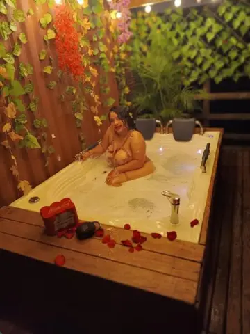 bigtits00022, a woman with dark hair, poses naked in a white bathtub filled with water and red rose petals, surrounded by green leaves and red flowers.