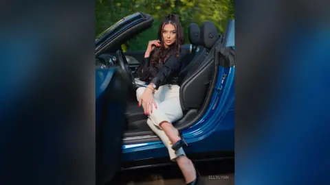 Stylish woman in black top and white trousers sitting in a blue convertible car outdoors.