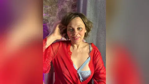 Smiling woman in a red lace robe and blue lingerie relaxing in a cozy, dimly lit room.
