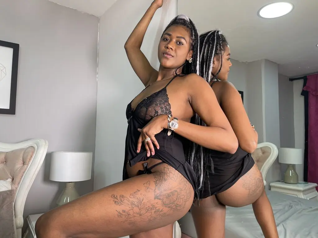 Confident woman in black lace lingerie posing in a bedroom mirror with floral leg tattoos.