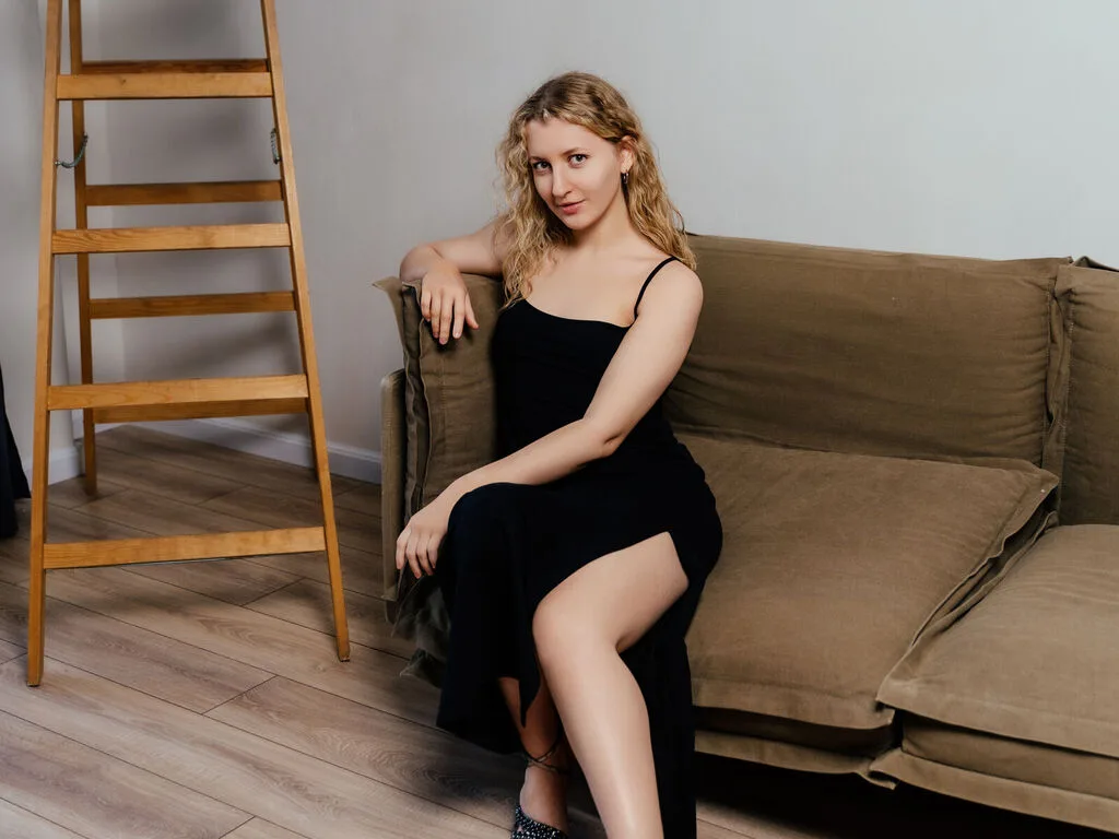 JolynJoestar sits on a brown sofa wearing a black slip dress with a high slit, showing her bare legs.