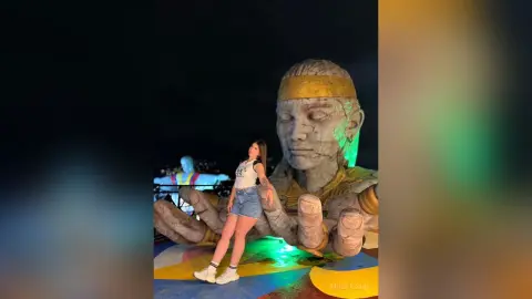 MiaBolson — Young woman in a white t-shirt and denim shorts standing before a giant stone Buddha statue at night.