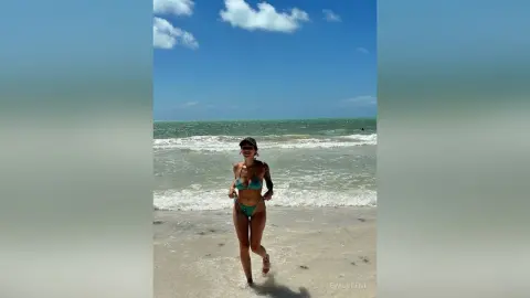 Young woman in a green bikini walking on a sunny beach with ocean waves under a blue sky.