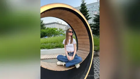Young woman in white t-shirt and jeans sitting on a circular wooden bench outdoors in a sunny urban park.