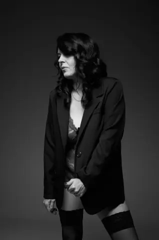 Moody black and white portrait of a woman in a dark blazer and lace lingerie against a gray studio background.