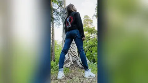 Young woman in black sweatshirt and blue jeans posing by a large tree in a sunny forest setting.