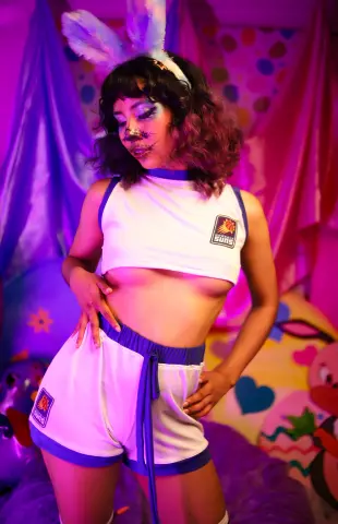 Woman in Phoenix Suns crop top and shorts cosplay outfit posing in a dreamy pink and purple lit room.