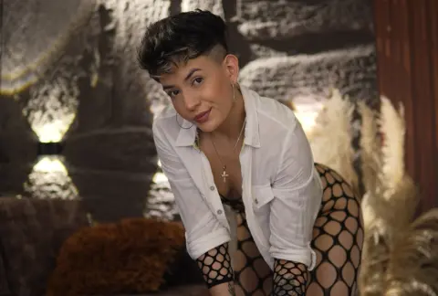 Smiling woman in white shirt and fishnet tights posing in a cozy, dimly lit room with warm lighting.