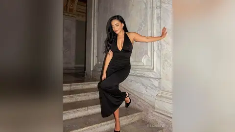 Elegant woman in a sleek black halter gown posing on marble stairs in a luxurious grand hallway.