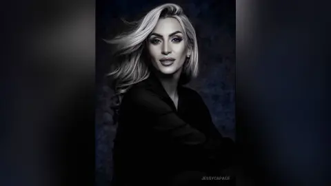 JessycaPage poses with her long blonde hair flowing against a dark background, wearing a black top.