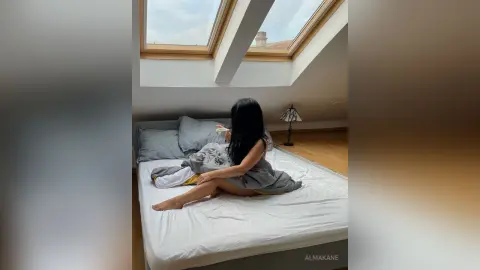 AlmaKane — Young woman in casual loungewear relaxing on a white bed in a bright, sunlit modern bedroom.