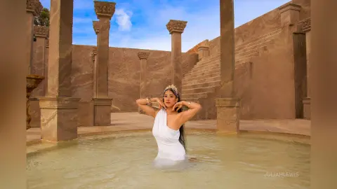 JuliahLewis poses in a white dress while waist-deep in a stone pool surrounded by classical columns.