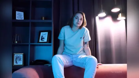 Young woman in a light blue t-shirt and jeans sitting on a couch in a moody, dimly lit room.
